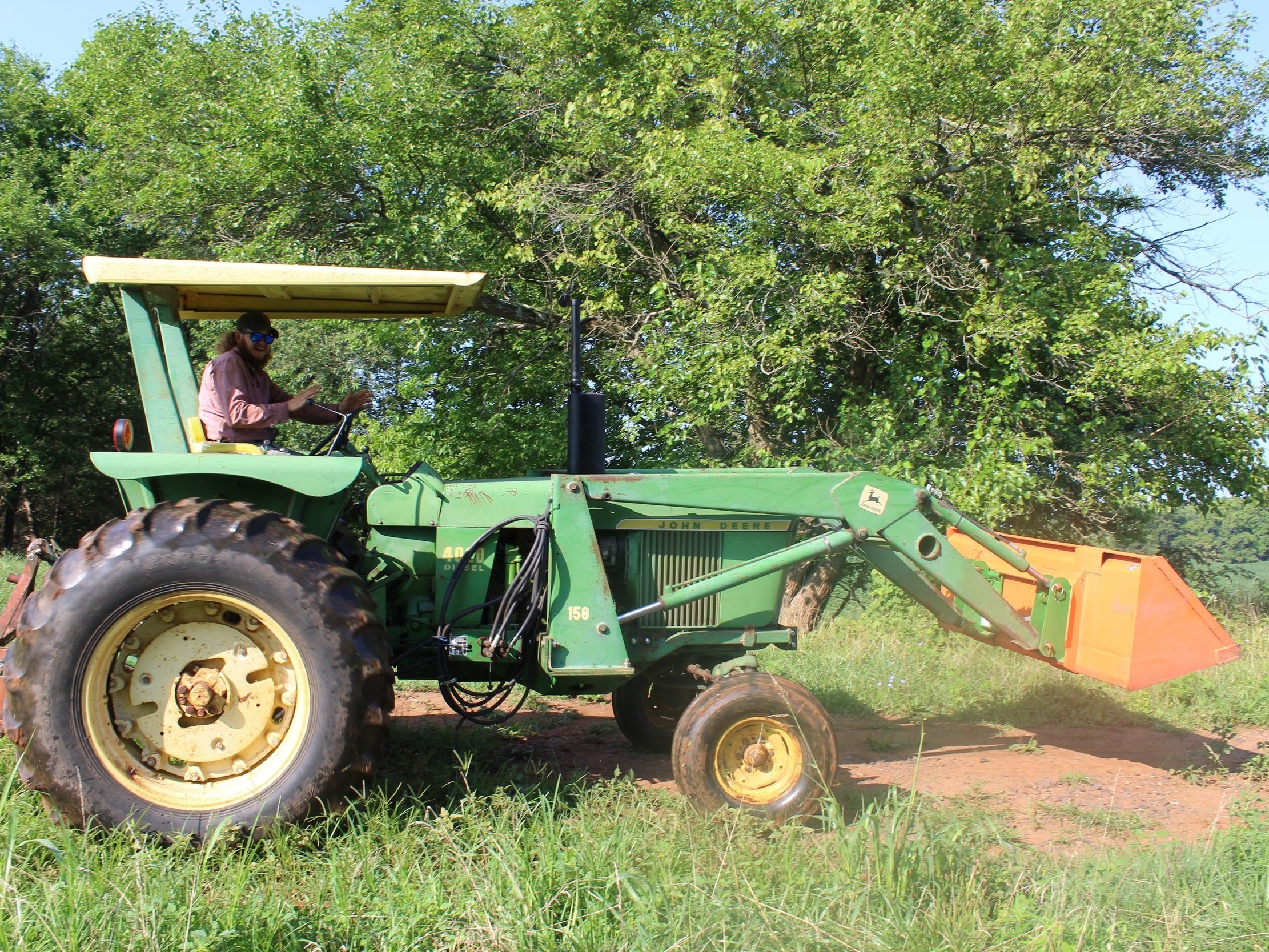 A farmer driving a green John Deere tractor with a front loader attachment, surrounded by lush greenery and grass. The tractor is parked on a dirt path, with the operator smiling and wearing sunglasses.