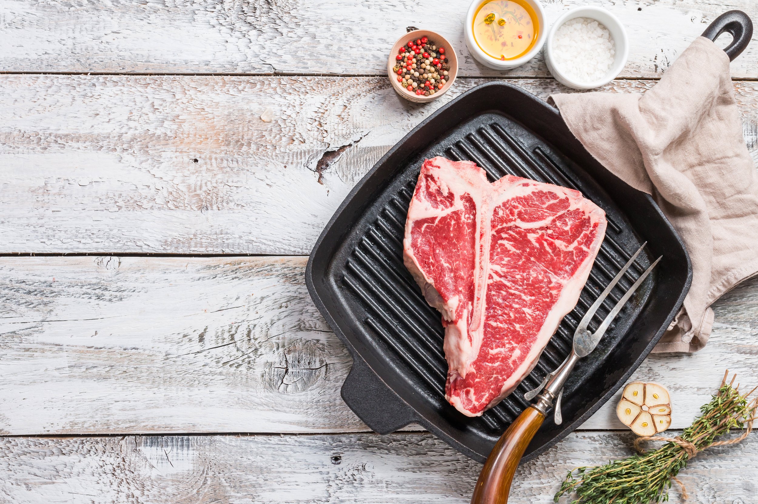 Raw T-bone steak on a grill pan with cooking utensils, surrounded by bowls of spices, olive oil, and fresh herbs on a rustic wooden surface.