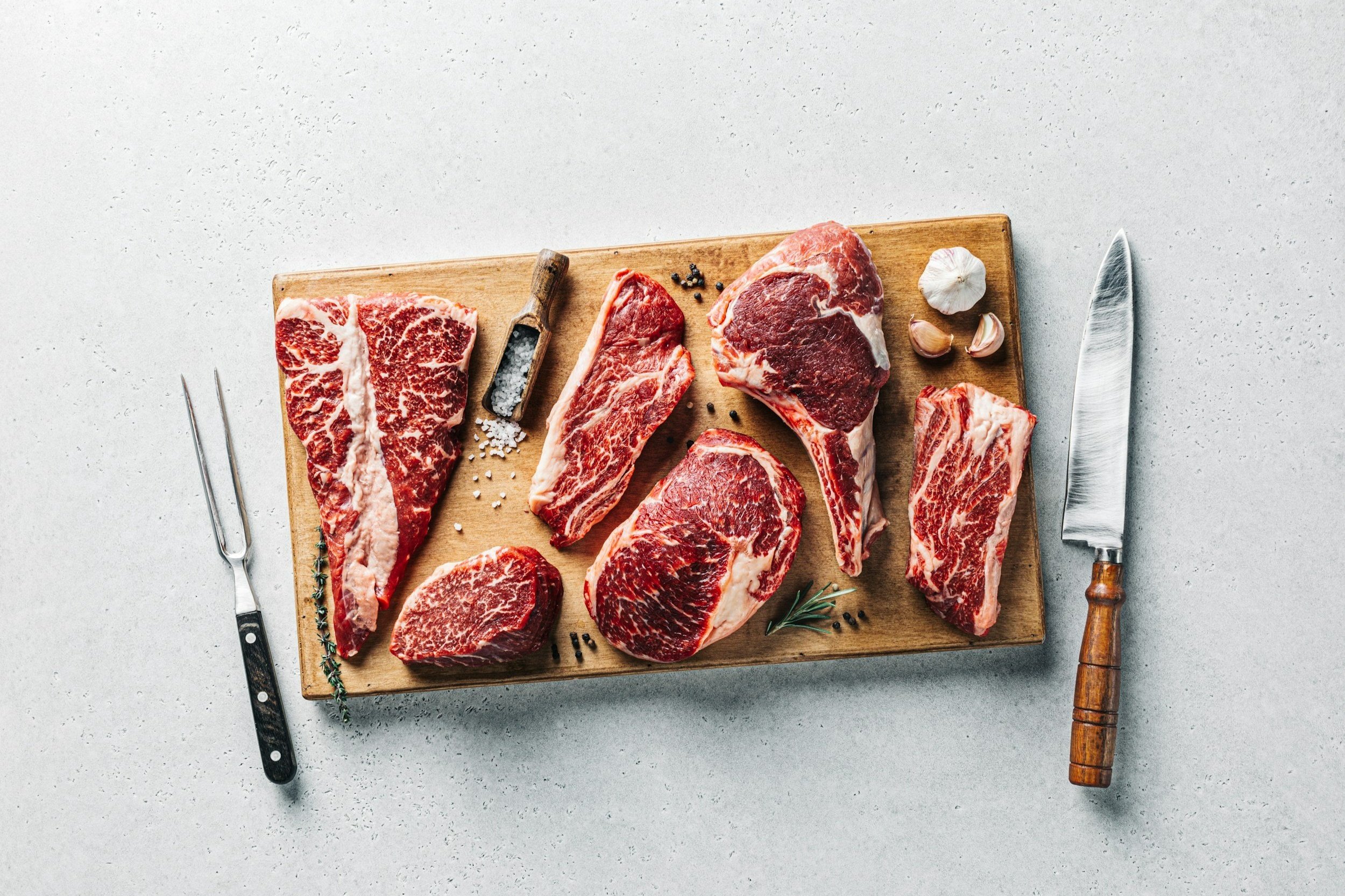 A wooden cutting board displaying various cuts of marbled beef, including ribeye and sirloin, accompanied by garlic cloves, sea salt, and a sharp knife, ideal for meat preparation and grilling.